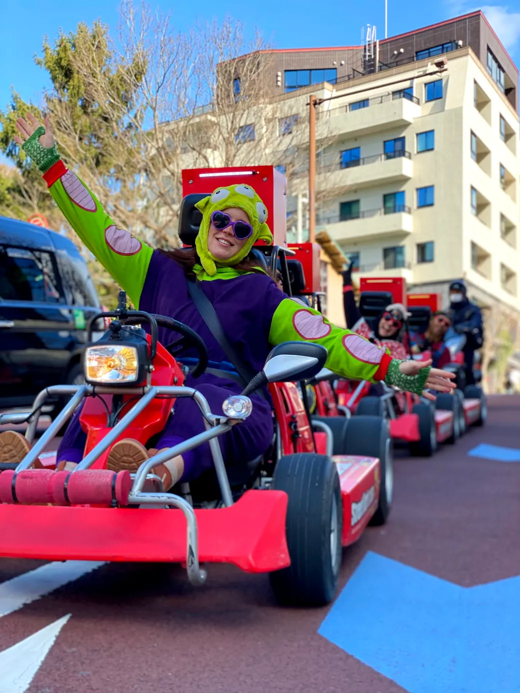 Person in a bright green and purple costume with an alien hat drives a red go-kart in a parade, waving happily. Behind them, other riders in colorful outfits follow in line on go-karts.