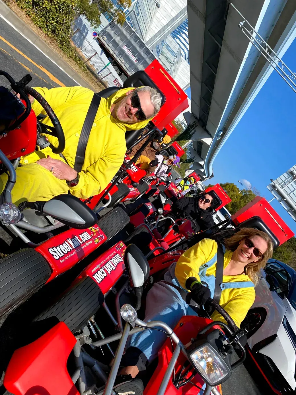 Group of people in bright yellow jackets riding red pedal go-karts along an urban street parade.