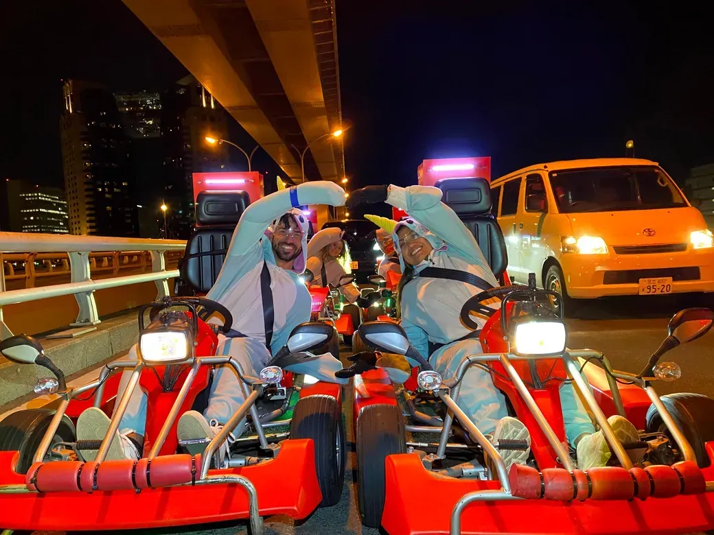 Two people in red go-karts on a city bridge at night form a heart shape with their arms, wearing white outfits and helmets, with a van in the background.