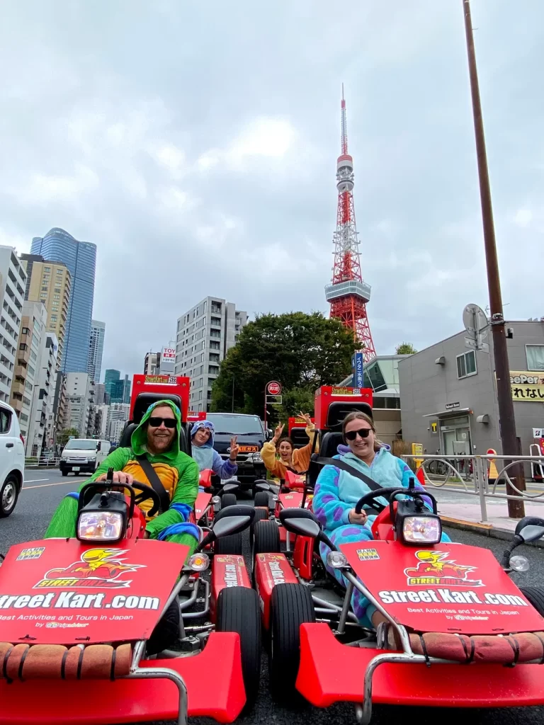 IMG_6165 - StreetKart Group of people in red go-karts with StreetKart logos driving on a city street, Tokyo Tower visible in the background, smiling faces.